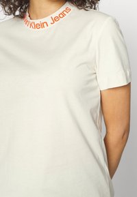 Close-up of a person wearing a white t-shirt with "Calvin Klein Jeans" in orange text around the neckline.