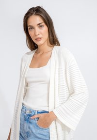Cardigan bianco a righe con design aperto, texture a costine e maniche larghe, indossato sopra un top a coste bianco e jeans blu.