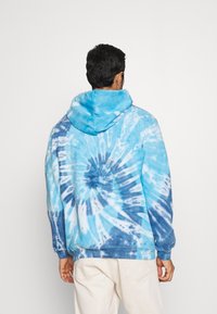 Uomo che indossa un hoodie tie-dye blu e bianco e pantaloni beige, visto da dietro su uno sfondo bianco semplice.