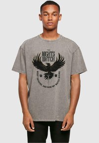 Grijze t-shirt met korte mouwen met een afbeelding van een adelaar en de tekst "DE NACHTWACHT" en "DE NACHT VERZAMELT EN NU BEGINT MIJN WACHT."