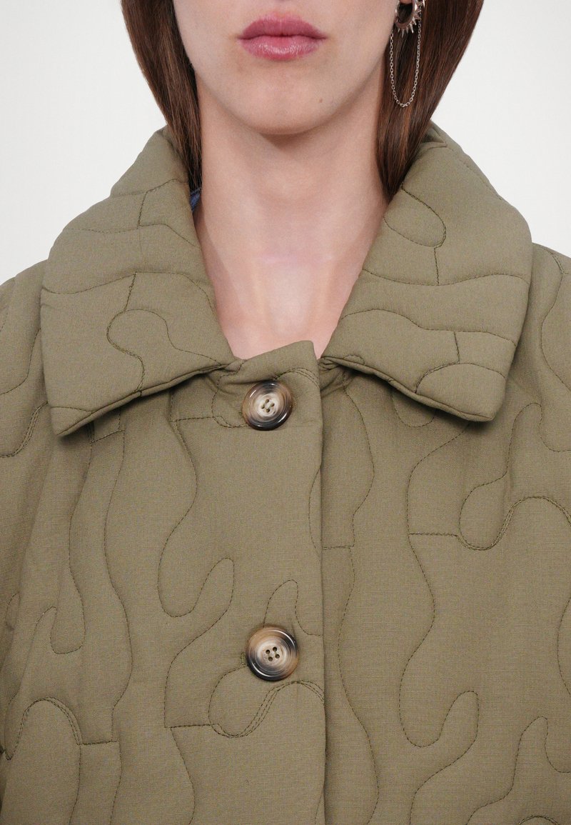 Veste matelassée vert olive avec un grand col et une texture lisse. Deux fermetures à bouton avec un motif subtil sur le tissu.