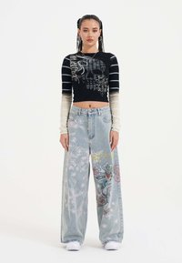Černý krátký top s grafickým designem; světle modré široké džíny s květinovými vzory. Modelka má na sobě bílé tenisky. Jednoduché pozadí.
