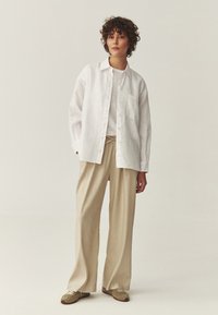 Mujer con cabello rizado que lleva una camisa blanca suelta abotonada, camiseta blanca, pantalones amplios beige y zapatillas beige, posando contra un fondo liso.