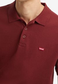 Polo bordeaux en tissu texturé, avec un col, quatre boutons et un petit logo Levi's rouge sur la poitrine.