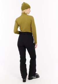 Persoon met een olijfgroene beanie en een lange mouw top, zwarte sneeuwbroek en zwarte skiboots, staat iets zijwaarts gekeerd op een witte achtergrond.