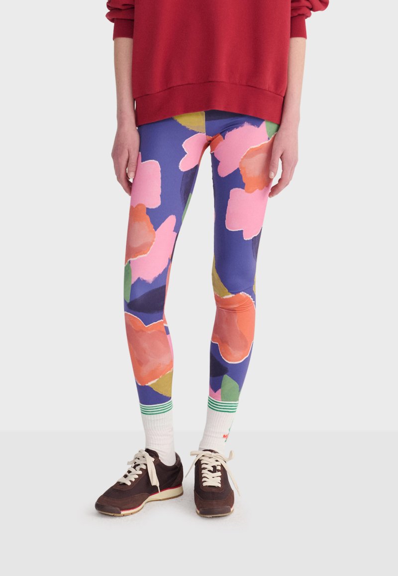 Jambes portant des leggings colorés avec de grands motifs floraux roses et orange, des chaussettes blanches à rayures vertes, et des baskets marron foncé.