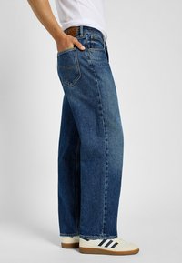 Jeans de mezclilla azul con un ajuste de pierna recta. Tela resistente, diseño de cinco bolsillos y costuras visibles. Combinados con zapatillas blancas.