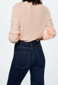 Hellrosa gestrickter Pullover mit langen Ärmeln, kombiniert mit hoch taillierter, dunkelblauer Denim-Jeans mit sichtbaren Gesäßtaschen und Nahtdetails.