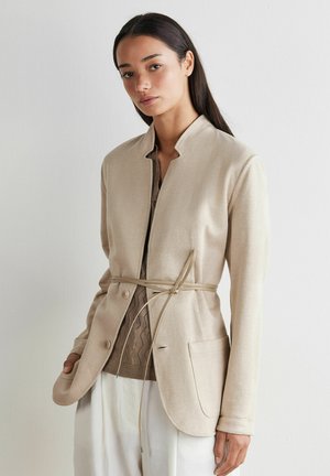 Frau trägt eine beige Jacke mit Knöpfen und einem dünnen Gürtel, der in der Taille gebunden ist, über einem braunen Strickoberteil und weißen Hosen, stehend vor einer weißen Wand.