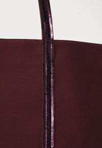 Bordeauxfarbene Tragetasche aus strukturiertem Stoff, die an einer Seite von einer vertikalen Linie glänzender, lila Pailletten akzentuiert wird.