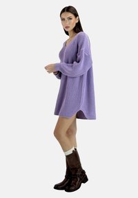Lila gestricktes Sweatkleid mit V-Ausschnitt, langen Ärmeln und gerippter Textur, kombiniert mit braunen kniehohen Stiefeln und weißen Socken.
