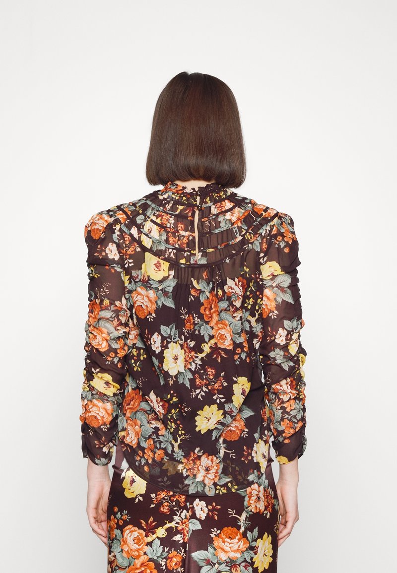Blouse transparente à fleurs sombres avec col à volants et manches longues. Présente un mélange de fleurs orange, jaune et verte sur un fond marron.