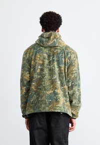 Hanorac din fleece cu un model de camuflaj în verde, maro și bej. Are glugă cu șnur și o croială lejeră. Textură moale.
