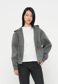 Pilkas užtrauktukų džemperis su tekstūriniu paviršiumi, reguliuojama kapuce, priekinėmis kišenėmis, derinamas su balta marškele ir tamsiomis džinsinėmis kelnių.