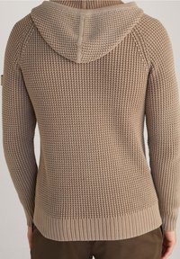Beige Waffelstrick-Hoodie, getragen über braunen Hosen, zeigt von hinten die detaillierte Strickstruktur und den gerippten Saum.