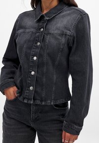 Veste en denim noir avec un col chemise, des boutons sur le devant et une coupe ajustée. Présente une texture légèrement usée et des détails de coutures.
