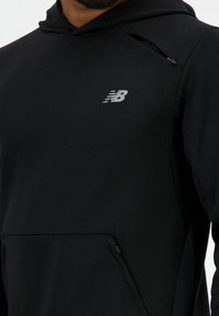 Sudadera negra con un bolsillo frontal que tiene una pequeña cremallera, hecha de un tejido suave y elástico. El logo de New Balance está en gris en el pecho.