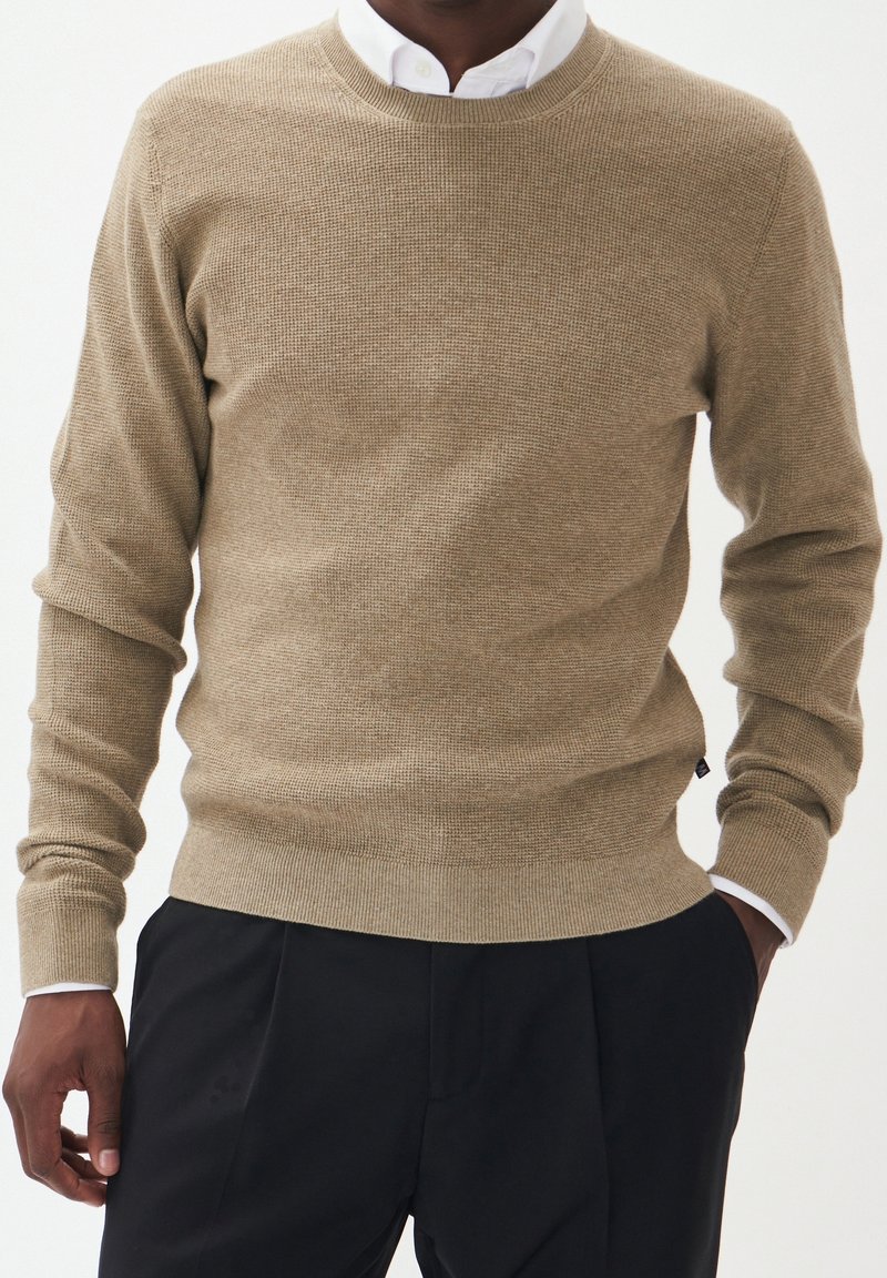 Homme portant un pull beige texturé par-dessus une chemise blanche à col et un pantalon noir, mains détendues le long du corps sur fond blanc.