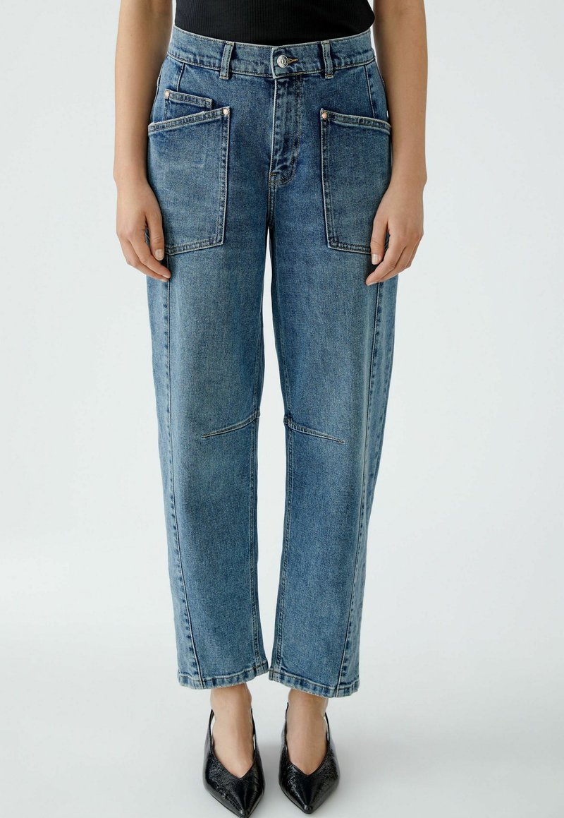 Frau in hochgeschnittenen blauen Jeans mit großen vorderen Patten-Taschen und schwarzen spitzen Absätzen, stehend vor einem schlichten Hintergrund.