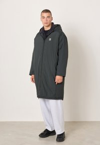 Muž v čiernom predĺženom páperovom kabáte s kapucňou, svetlosivých teplákoch a čiernych teniskách stojaci na neutrálnom pozadí.