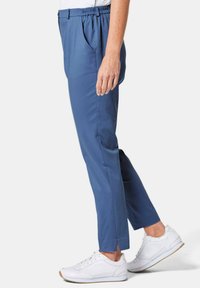 Pantalon bleu sur mesure avec une taille élastique, des poches latérales et des plis subtils, associés à des baskets blanches. Les pantalons ont des fentes aux chevilles.