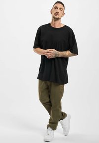 Maglietta nera oversize, pantaloni verde oliva larghi e sneakers bianchi. Braccia tatuate visibili, sfondo neutro.