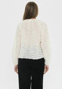 Donna con lunghi capelli castani che indossa un top bianco trasparente a maniche lunghe e pantaloni neri, rivolta di spalle su uno sfondo uniforme.