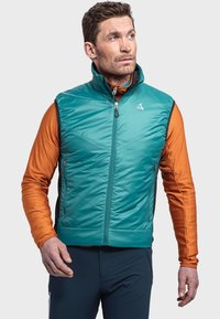 Türkisfarbene, isolierte Weste mit hohem Kragen und Frontreißverschluss, getragen über einem orangefarbenen Langarmshirt mit einem dezenten Chevron-Muster und mit Reißverschlusstaschen.