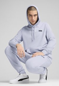 Lyslilla hoodie og sweatpants sæt lavet af blødt stof, med en kængurulomme og Puma-logo, parret med hvide sko med sorte accenter.