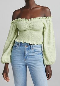 Top verde chiaro senza spalline con texture arricciata, bordi volant e maniche lunghe a palloncino, abbinato a jeans blu a vita alta.