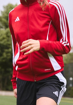 Veste de sport rouge zippée avec des rayures blanches, fabriquée en tissu léger, associée à un short noir avec des accents blancs et une coupe athlétique.