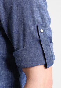 Oprullet ærme på en blå struktureret skjorte med knapper, der viser armen og stofdetaljerne.