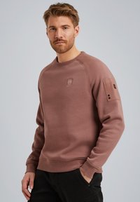 Sudadera color burdeos con cuello redondo, puños acanalados y un discreto parche con el logo en el pecho. Presenta un bolsillo en la manga.