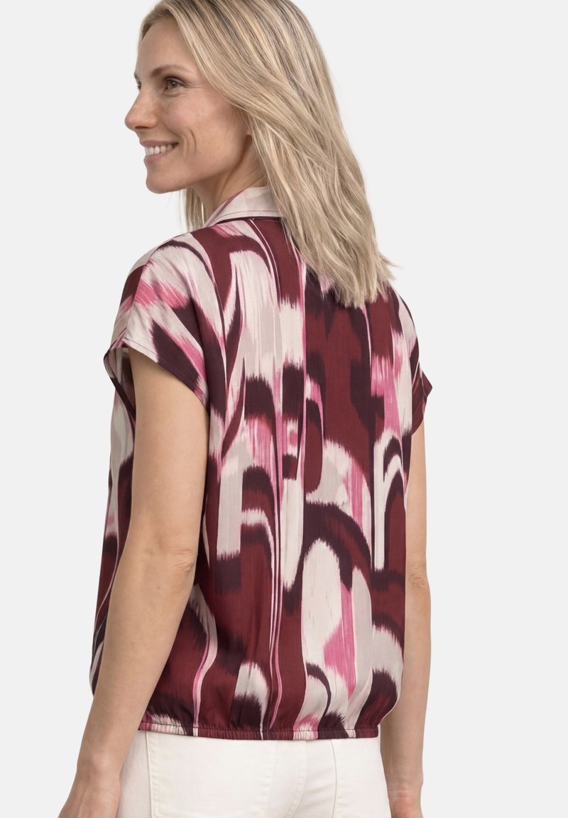 Femme aux cheveux blonds sourit, portant un chemisier à manches courtes avec des motifs abstraits bordeaux, rose et crème, associé à un pantalon blanc.