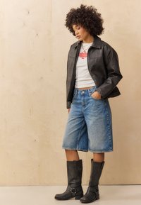 Veste en cuir noire, t-shirt blanc à motif graphique, shorts en denim bermuda et bottes noires jusqu'au genou avec des accents de boucle. Cheveux bouclés. Fond neutre.