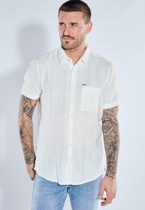 Mann mit kurzen Haaren und Tattoos an beiden Armen, trägt ein weißes kurzärmeliges Hemd und hellblaue Jeans, schaut zur Seite.