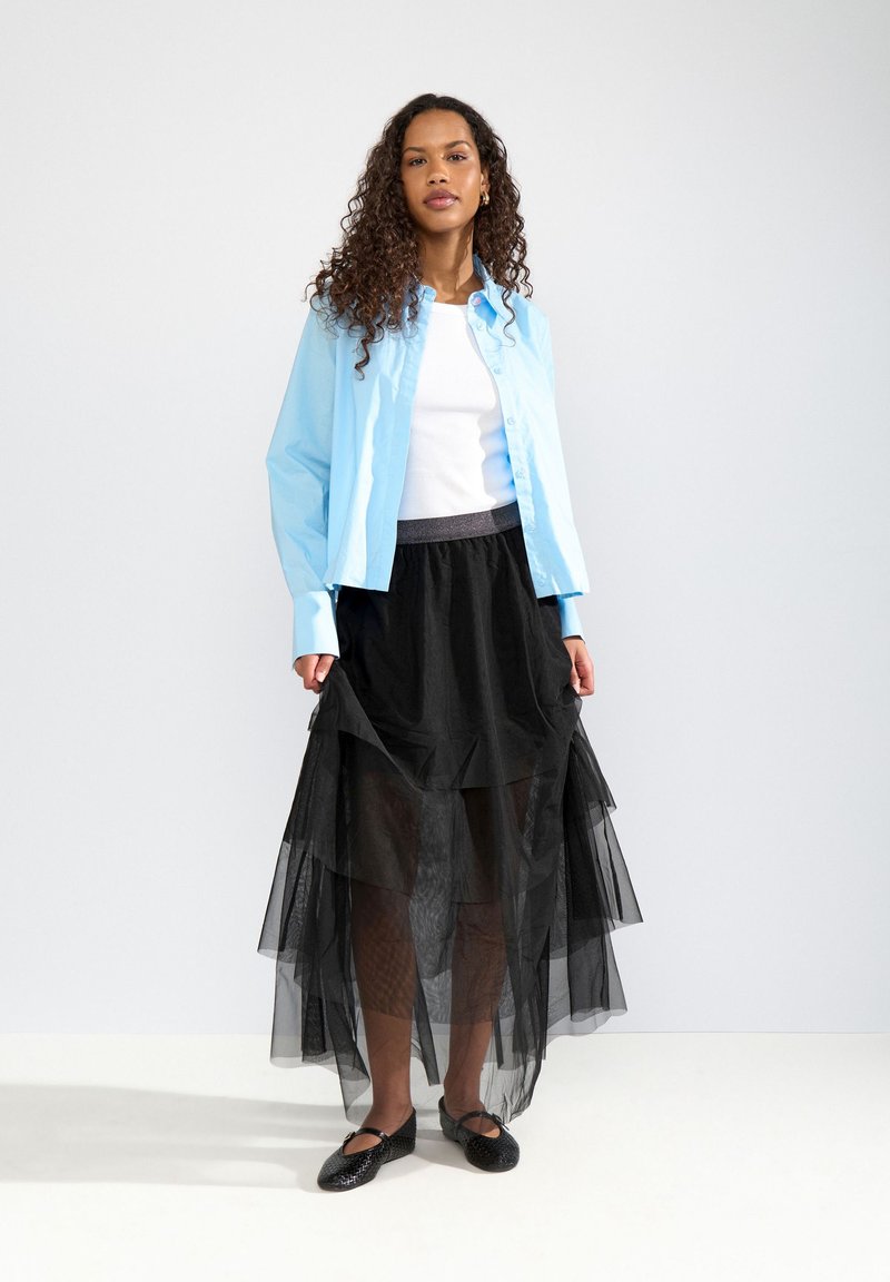 Femme aux cheveux bouclés portant une chemise bleu clair boutonnée sur un haut blanc, une jupe en maille noire à plusieurs couches et des ballerines noires, debout sur un sol blanc.