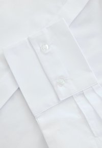 Bílá košile s límcem a dvěma knoflíkovými zapínáními, hladká tkanina a skládací část, která ukazuje čistý a upravený design.