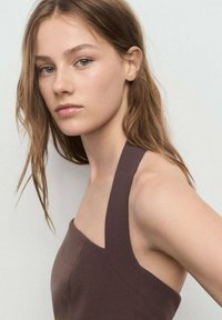 Vestido marrón de cuello halter con tejido suave, diseño ajustado y costuras mínimas. El cabello es largo y ondulado, con un fondo neutro.