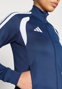 Αθλητικό μπουφάν σε ναυτικό μπλε απόχρωση με ψηλό γιακά, λευκές λωρίδες στα πλαίσια των ώμων και φερμουάρ μπροστά. Διαθέτει υφασμάτινη υφή και πλαϊνές τσέπες.