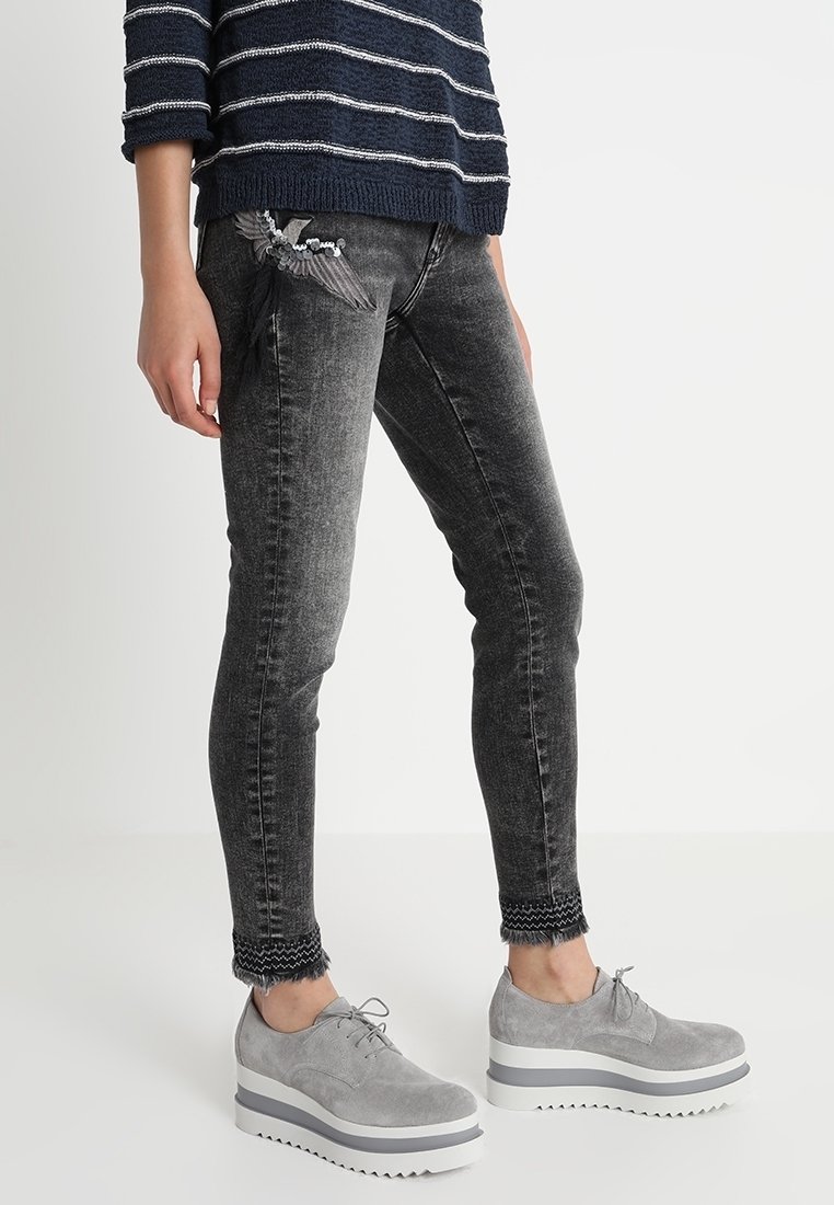 Jeans ajustados grises con detalle bordado de un pájaro en el bolsillo, dobladillo deshilachado, combinados con zapatos de plataforma grises y un suéter a rayas azul marino.