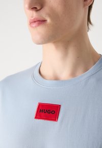 Tricou din bumbac de culoare albastru deschis, cu guler rotund; are o aplicație rectangulară roșie cu cuvântul "HUGO" în negru, plasată pe piept.