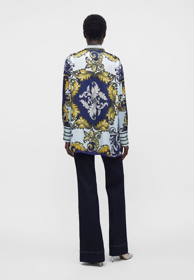 Personne portant une chemise à manches longues avec des motifs baroques bleus, blancs, jaunes et dorés, associée à un pantalon ample foncé et des talons blancs.