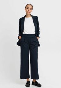 Blazer blu navy su una camicia bianca, abbinato a pantaloni larghi a vita alta blu navy. Tessuto testurizzato e linee pulite con tasche laterali. Mocassini neri.