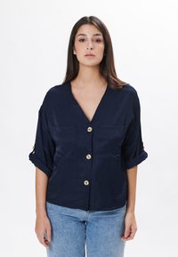 Marineblaue Bluse mit V-Ausschnitt, zwei vorderen Taschen und goldenen Knopfakzenten. Verfügt über aufgerollte Ärmel und eine lockere Passform. Glatte Textur.