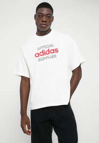 Mladenič oblečen v belo majico s tekstom "Uradni dobavitelj adidas" v rdeči in črni barvi, v kombinaciji s črnimi hlačami na enobarvnem ozadju.