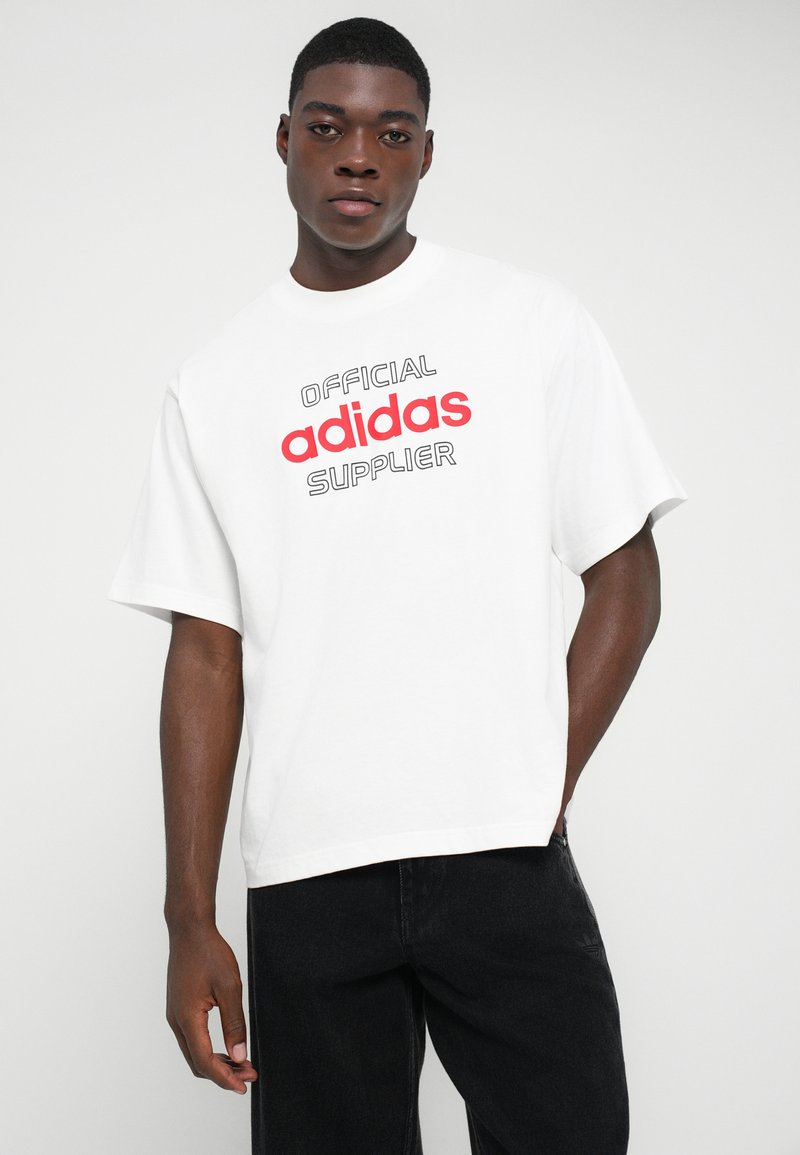 Joven vistiendo una camiseta blanca con el texto "Proveedor Oficial de adidas" en rojo y negro, combinada con pantalones negros frente a un fondo sencillo.