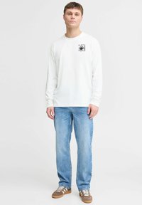 Camiseta blanca de manga larga con un estampado gráfico negro en el pecho, combinada con jeans de mezclilla azul claro y zapatillas marrones.