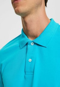 Polo turquoise avec une finition texturée, doté d'un col classique et d'une patte à deux boutons avec des boutons bleu clair.