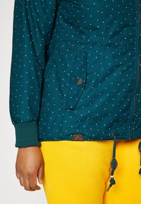 Teal jacket with colorful polka dots, elastic cuffs, and adjustable drawstring. Features a front pocket and a brand tag on the hem.